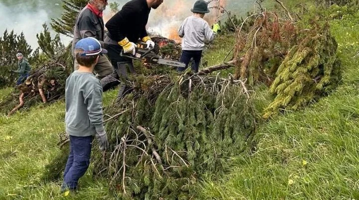 Auf der Bergwiese liegen Baum- und Strauchabschnitte. Im Hintergrund sieht man, wie ein Teil verbrannt wird, im Vordergrund fleißige Helfer | © Sebastian Pertl